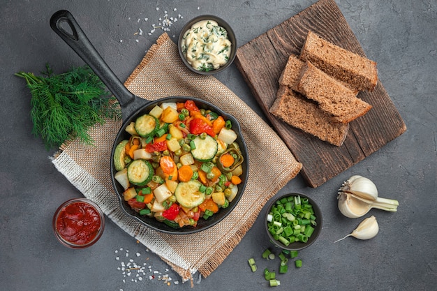 Gesundes Mittagessen mit Gemüseeintopf, frischen Kräutern und Roggenbrot auf graubraunem Hintergrund. Ansicht von oben, horizontal.