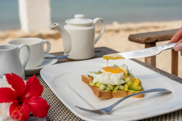 Gesundes Sandwich mit Avocado, Gurke und pochierten Eiern auf dem Tisch für gesundes Frühstück am Strand in der Nähe des Meeres. Food and Breakfast-Konzept.