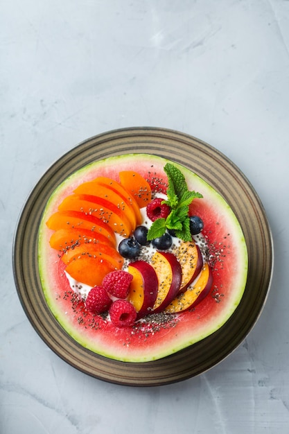 Gesundes sauberes Essen Ernährung und Ernährung saisonales Sommerfrühstückskonzept Obstsalat mit Joghurt in geschnitzter Wassermelonenschüssel auf einem Küchentisch Draufsicht flach liegend Kopierraum Hintergrund