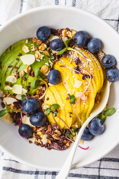 Gesundes veganes Frühstück. Granola mit Avocado, superfoods, Beeren und Früchten in einer weißen Schüssel, Draufsicht.