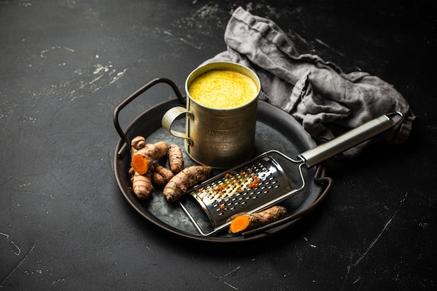 Gesundes Warmgetränk zur Stärkung des Immunsystems, Kurkuma-Goldmilch. Zutaten zum Kochen von Detox-Curcuma-Milch in rustikaler Zinnschale, frische Kurkumawurzel, Reibe auf schwarzem Steinhintergrundkonzept