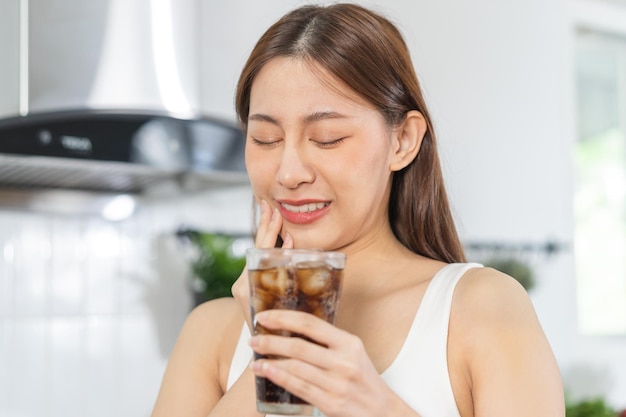 Gesundheit asiatische junge frau, die den wangenausdruck berührt, der an zahnschmerzen, verfall oder empfindlichkeitshöhlenzahnzähnen oder entzündungen leidet, trinkt kaltes sprudelndes wasser zu hause. Menschen mit empfindlichen zähnen