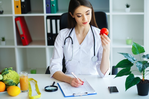 Gesundheit. Der Arzt stellt einen Diätplan auf. Der Ernährungsberater hält in den Händen von frischen Tomaten. Früchte und Gemüse. Junger Doktor mit einem schönen Lächeln im Büro.