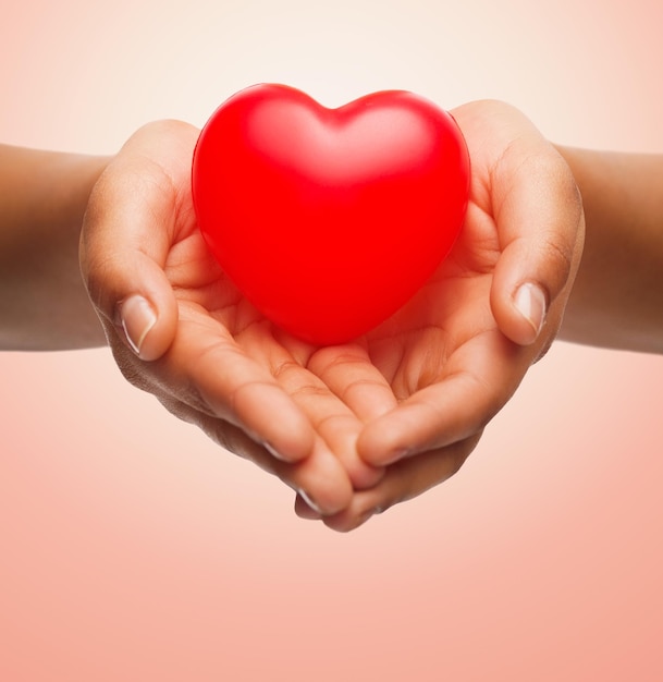 Gesundheit, Medizin, Liebe, Valentinstag und Wohltätigkeitskonzept - Nahaufnahme von afroamerikanischen Frauenhänden, die ein kleines rotes Herz über beigem Hintergrund halten