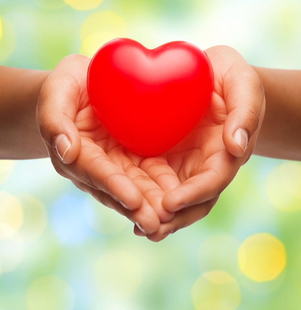 Gesundheit, Medizin, Liebe, Valentinstag und Wohltätigkeitskonzept - Nahaufnahme von afroamerikanischen weiblichen Händen, die ein kleines rotes Herz über grünem Hintergrund halten