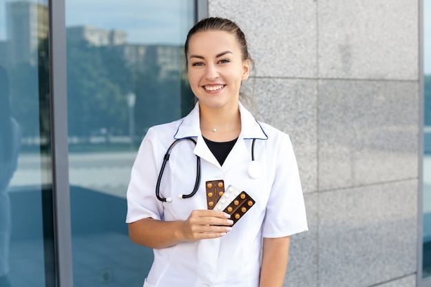 Gesundheits-, Berufs- und Menschenkonzept - Europäische lächelnde Ärztin in einem weißen Kittel mit einem Stethoskop, das mehrere Pillenpakete hält und bei gutem Wetter draußen in die Kamera schaut