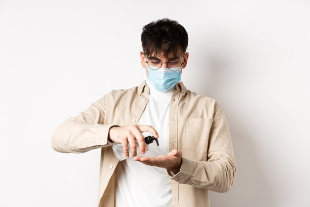 Gesundheits-, Covid- und Quarantänekonzept. Junger Hispanic Kerl in Brille und Gesichtsmaske mit Händedesinfektionsmittel, Antiseptikum anwenden, auf weißer Wand stehend.