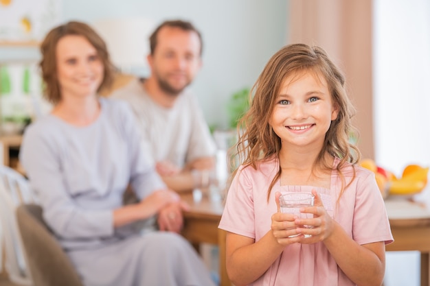 Gesundheits- und Schönheitskonzept - lächelndes kleines Mädchen, das ein Glas Wasser hält. Glückliche Eltern im Hintergrund.