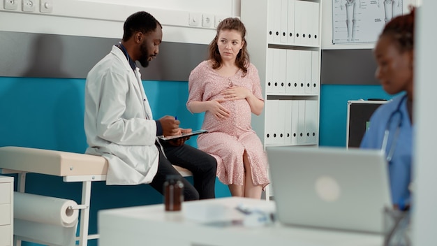 Gesundheitsspezialist, der eine Checkup-Beratung mit einem werdenden Patienten im Büro durchführt. Männlicher Arzt, der ein digitales Tablet verwendet, um sich Notizen zu machen und Frauen mit Schwangerschaftsbauch medizinischen Rat zu geben.