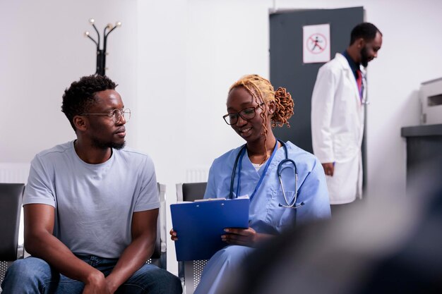 Gesundheitsspezialist untersucht jungen Mann in der Lobby des Wartebereichs und macht sich Notizen zu Berichtspapieren, um Medikamente mit Versicherungsunterstützung zu geben. Patient und Krankenschwester bei der Untersuchungsberatung im Wartezimmer.