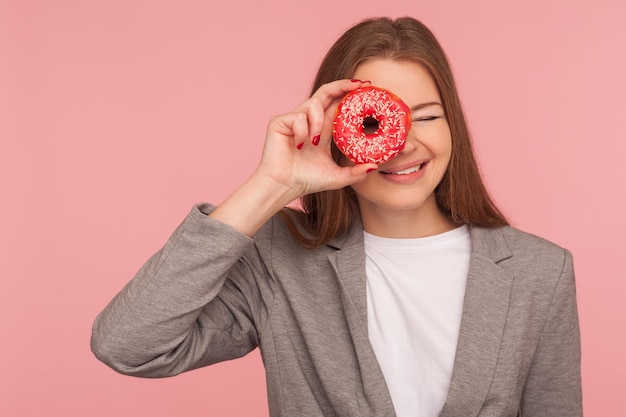 Gesundheitsvorsorge und Ernährung. Porträt einer fröhlichen Geschäftsfrau in Anzugjacke, die durch süßen Donut schaut, Spaß mit Gebäck hat, in Donut späht und lächelt. Studioaufnahme im Innenbereich, rosafarbener Hintergrund