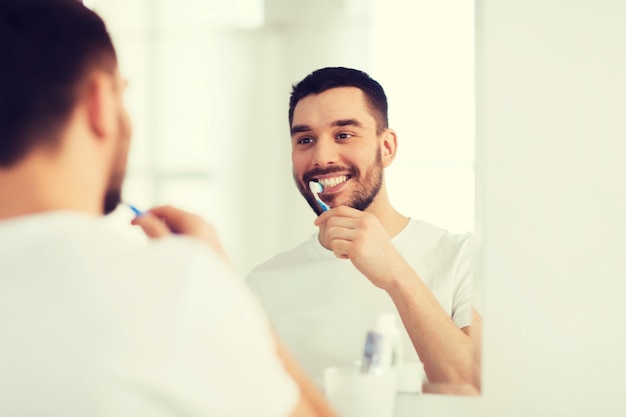 Gesundheitswesen, Zahnhygiene, Menschen und Schönheitskonzept - lächelnder junger Mann mit Zahnbürste, der die Zähne putzt und auf den Spiegel im heimischen Badezimmer blickt