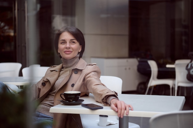 Getönte Lifestyle-Porträtfotografie einer attraktiven, atemberaubend schönen jungen eleganten Frau in stilvoller Freizeitkleidung, die in der Cafeteria im Freien ruht und eine Kaffeepause genießt