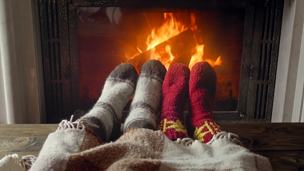 Getöntes Foto von Paarfüßen, die warme Socken tragen, die unter einer Decke am brennenden Kamin liegen