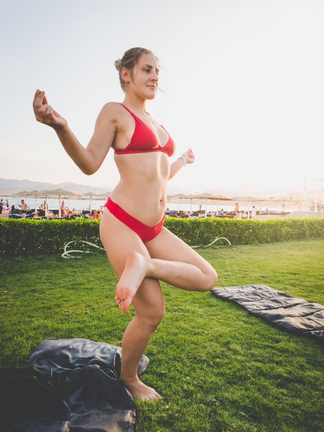 Getontes Bild einer jungen schlanken Frau mit rotem Bikini, die Yoga-Übungen auf dem Gras neben dem Meeresstrand macht