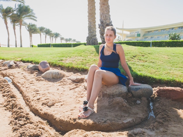 Getontes Foto einer schönen jungen Frau im kurzen Rock, die auf dem Felsen unter einer großen Palme am Meeresstrand sitzt