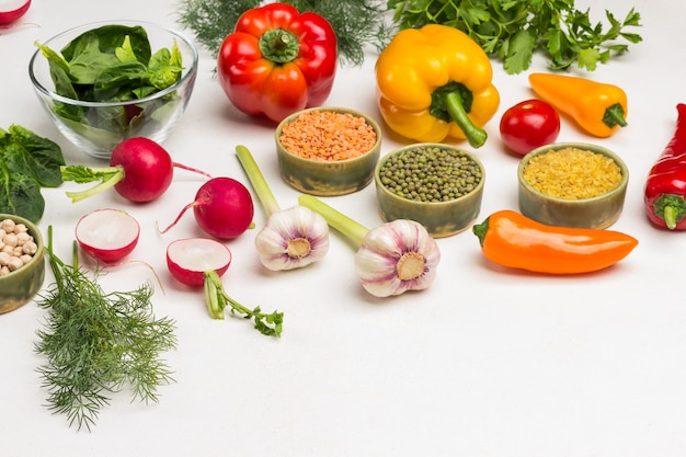 Getreide, Gemüse und Grüns auf dem Tisch. Püree, Linsen, Bulgur in Schalen. Radieschenhälften, Knoblauch und farbige Paprika. Weißer Hintergrund. Ansicht von oben. Platz kopieren