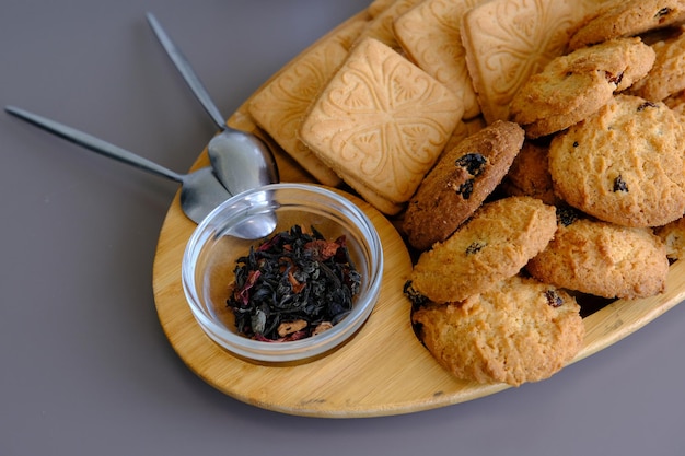 Getrocknete Teeblätter mit Teekanne und Plätzchen auf einem grauen Tisch mit Löffeln und Holzbrett