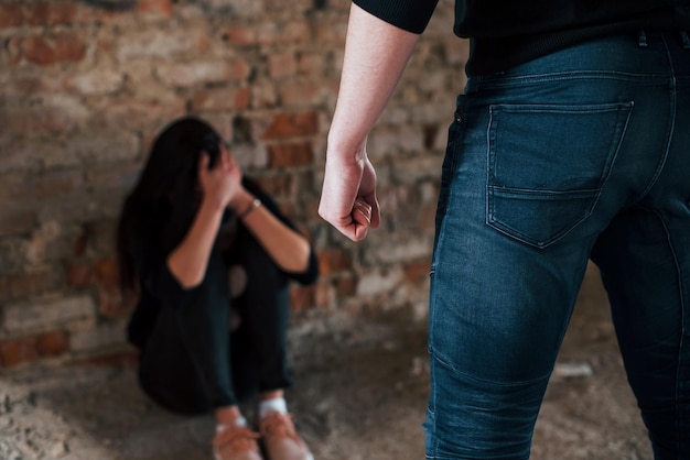 Gewalttätiger Mann steht und bedroht Mädchen, das mit Teddybär in verlassenem Gebäude auf dem Boden sitzt