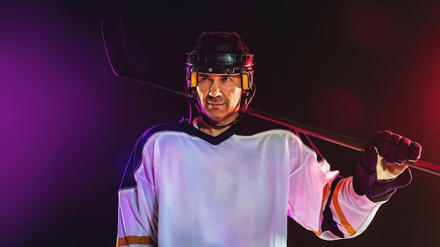 Gewinner. Männlicher Hockeyspieler mit dem Stock auf dem Eisplatz und der dunklen neonfarbenen Wand Sportler mit Ausrüstung, Helmübungen. Konzept des Sports, des gesunden Lebensstils, der Bewegung, des Wellness, der Aktion.