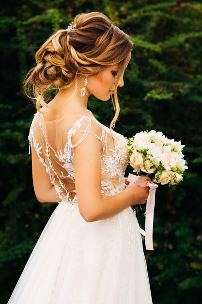 Gezeugte Braut in einem schönen Kleid mit einer bezaubernden Frisur hält einen Hochzeitsblumenstrauß