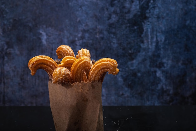 Gezuckerte Churros auf schwarzem Tisch mit Reflektion. Churros in Papiertüte. Blauer Hintergrund.