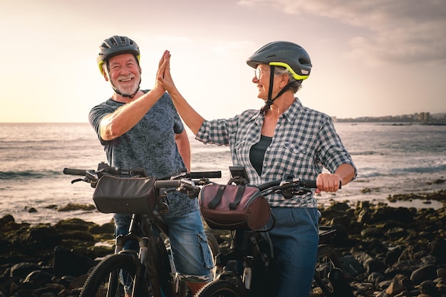 Gib mir fünf Älteres fröhliches Paar, das bei Sonnenuntergang mit Elektrofahrrädern am Kieselstrand reitet Authentisches älteres Leben im Ruhestand und nachhaltiges Mobilitätskonzept