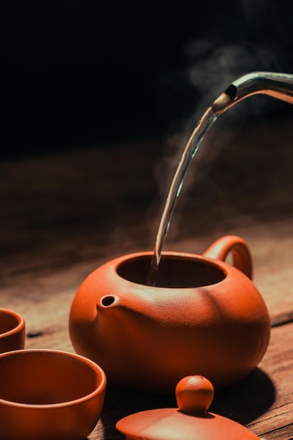 Gießen Sie heißes Wasser in einen Teekrug aus Ton für duftenden roten Rosentee Platziertes altes Holz mit schwarzem Hintergrund