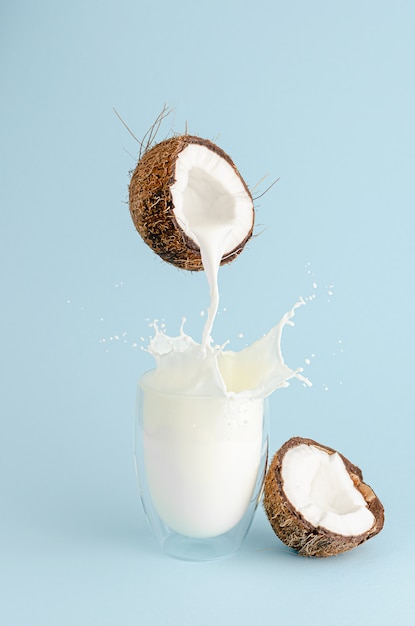 Gießen Sie Milch aus einer Kokosnuss und spritzen Sie in ein Glas auf pastellblauem Hintergrund. Konzept der Lebensmittelschwebung. Vertikale
