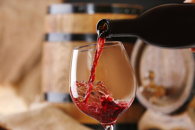 Gießen von Rotwein aus der Flasche in Glas mit hölzernen Weinfässern im Hintergrund