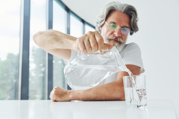 Gießt Wasser in Glas Älterer stilvoller moderner Mann mit grauem Haar und Bart drinnen