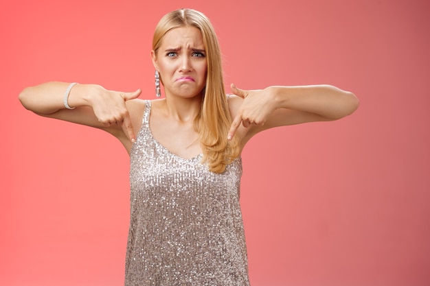 Gimiendo quejándose molesta mujer rubia deprimida en vestido plateado llorando novio rompió la noche de graduación de pie infeliz expresando tristeza arrepentimiento decepción apuntando hacia abajo de fondo rojo.