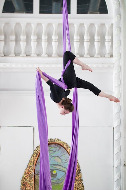 Gimnasta de aire joven en una ropa deportiva negra realiza un ejercicio sobre seda aérea en un estudio luminoso