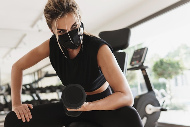 Ginásio novo normal. jovem mulher atlética usando uma máscara facial de prevenção durante seu treino de fitness com halteres.