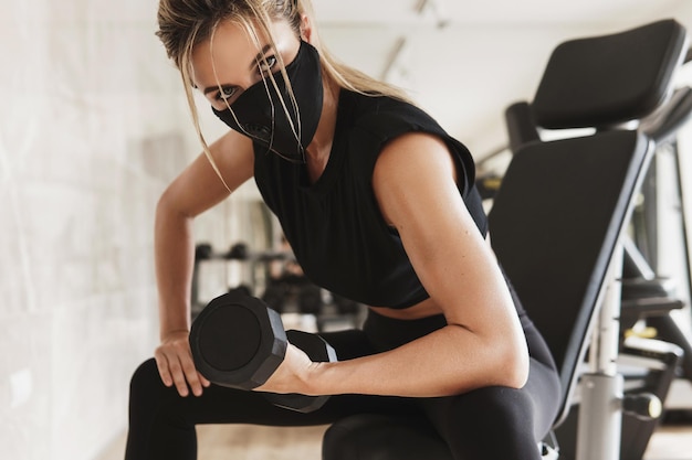 Ginásio novo normal. jovem mulher atlética usando uma máscara facial de prevenção durante seu treino de fitness com halteres.