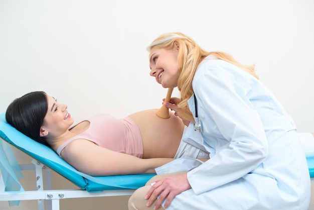 Ginecólogo obstetra escuchando el latido del corazón del feto infantil de una mujer embarazada feliz con