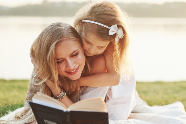 Girly tenta ler tão rápido. Mãe e filha lendo um livro em um dia ensolarado deitado na grama com o lago ao fundo.