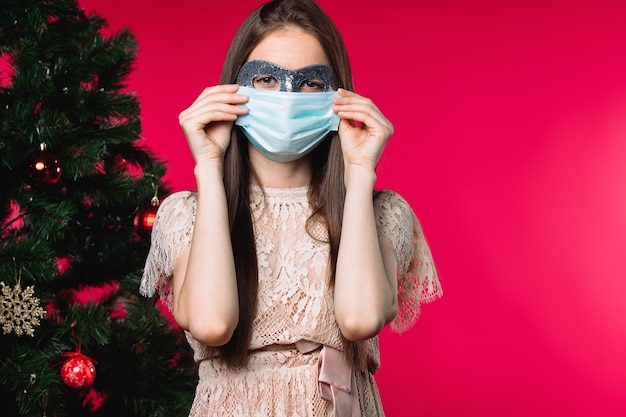 Girrl com carnaval e máscara médica em um fundo vermelho ao lado da árvore de natal