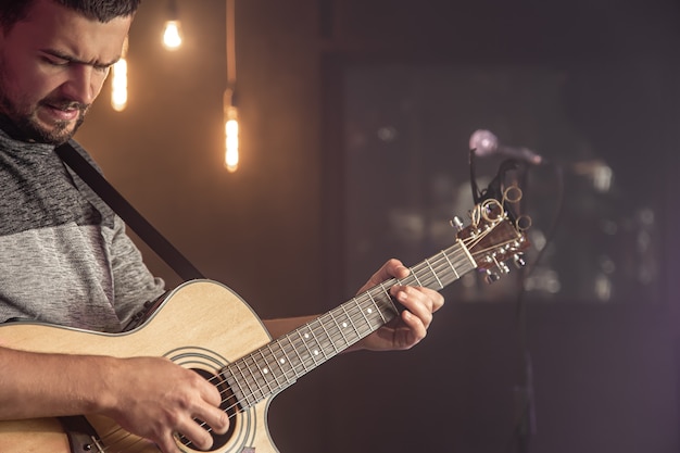 Gitarrist, der akustische Gitarre gegen unscharfen dunklen Hintergrund bei Konzertnahaufnahme spielt.