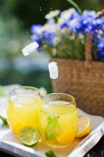 Gläser mit Limonade mit Minze, Limetten, Zitronen und Orange auf einem weißen Holztablett in der Nähe von Strohsack mit Wildblumen Sommergetränkekonzept