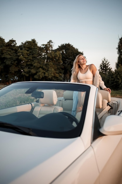 Una glamorosa chica de lujo con cabello rubio sonriendo mientras está sentada en un descapotable blanco. Joven mujer exitosa sentada en su auto blanco