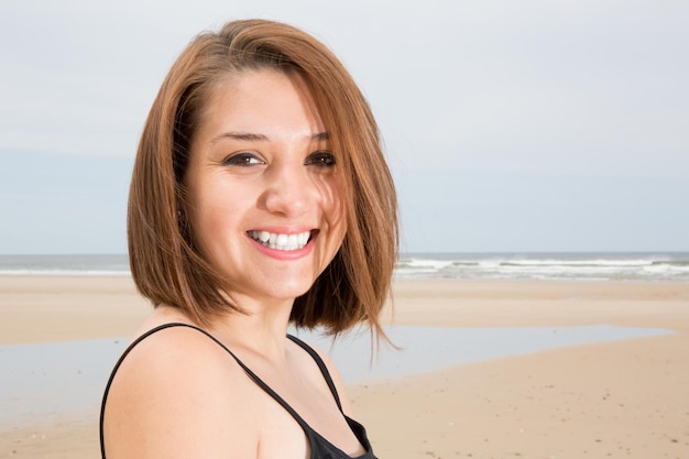 Glamorosa chica morena sonriente con vestido negro posando en la playa durante el atardecer