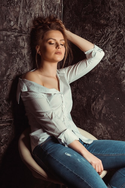 Glamorosa modelo joven sentada en una silla en jeans rayados y rasgados