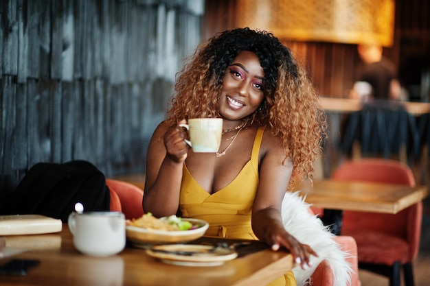 Glamour-Afroamerikanerin in gelbem Kleid, die mit einer Tasse Tee im Restaurant sitzt