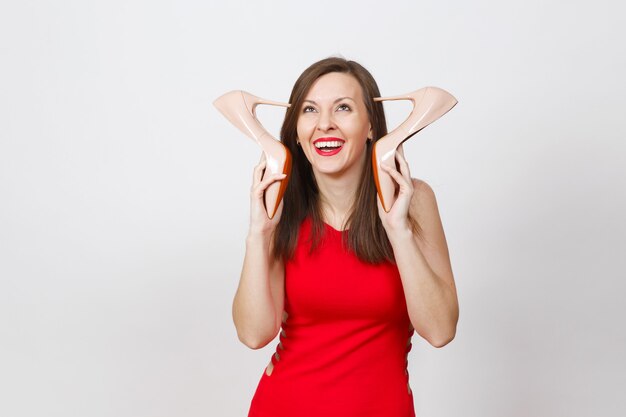 Glamour atraente na moda jovem de cabelos castanhos mulher vestida de vermelho, colocando os saltos altos na cabeça, ficando os sapatos bege com sola vermelha de suas compras isoladas no fundo branco. copie o espaço para anúncio.