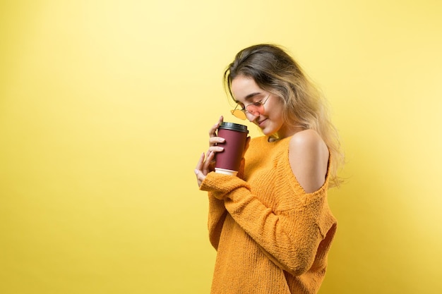 Glamour-Frau in Gläsern in einem orangefarbenen Pullover mit einem Drink Kaffee auf gelbem Hintergrund