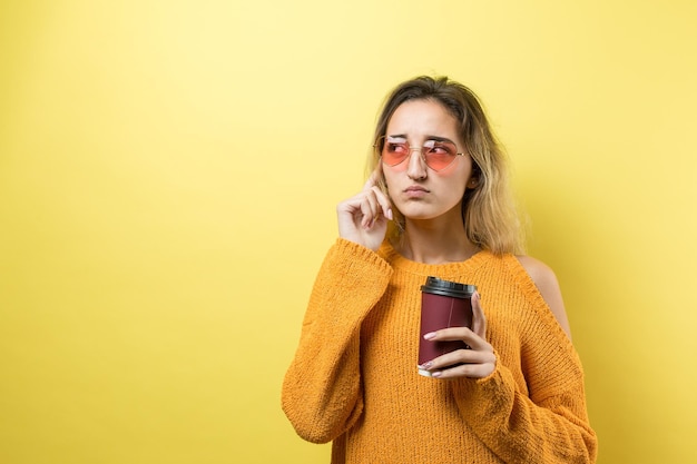 Glamour-Frau in Gläsern in einem orangefarbenen Pullover mit einem Drink Kaffee auf gelbem Hintergrund