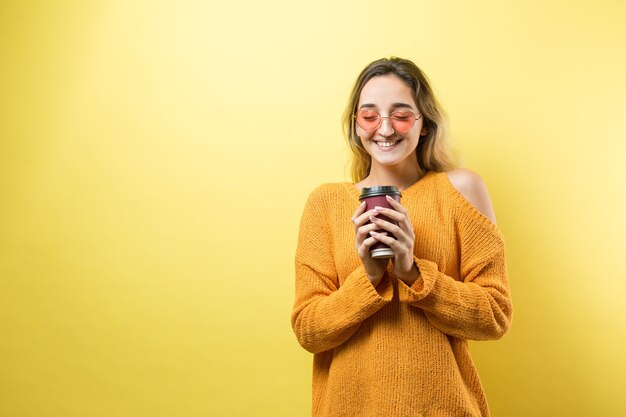 Glamour-Frau in Gläsern in einem orangefarbenen Pullover mit einem Getränk Kaffee auf gelbem Hintergrund