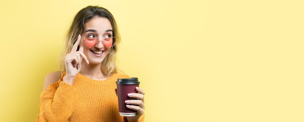 Glamour-Frau in Gläsern in einem orangefarbenen Pullover mit einem Getränk Kaffee auf gelbem Hintergrund