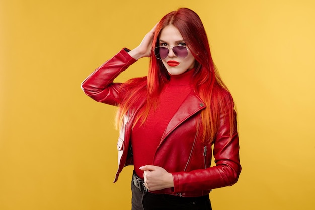 Glamour-Frau mit Sonnenbrille in roter Lederjacke und mit roten Haaren posiert auf Studiohintergrund